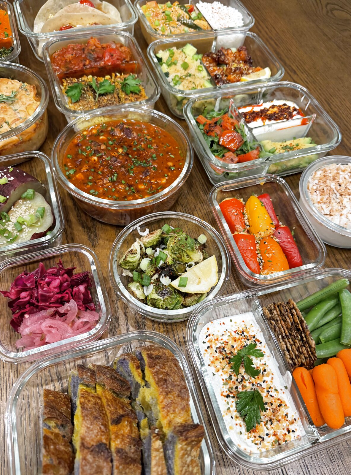 A spread of weekly meal prep in glass containers — stuffed mini peppers, roasted brussels sprouts, turkey chili, whipped feta with veggies, pickled cabbage, and shakshuka